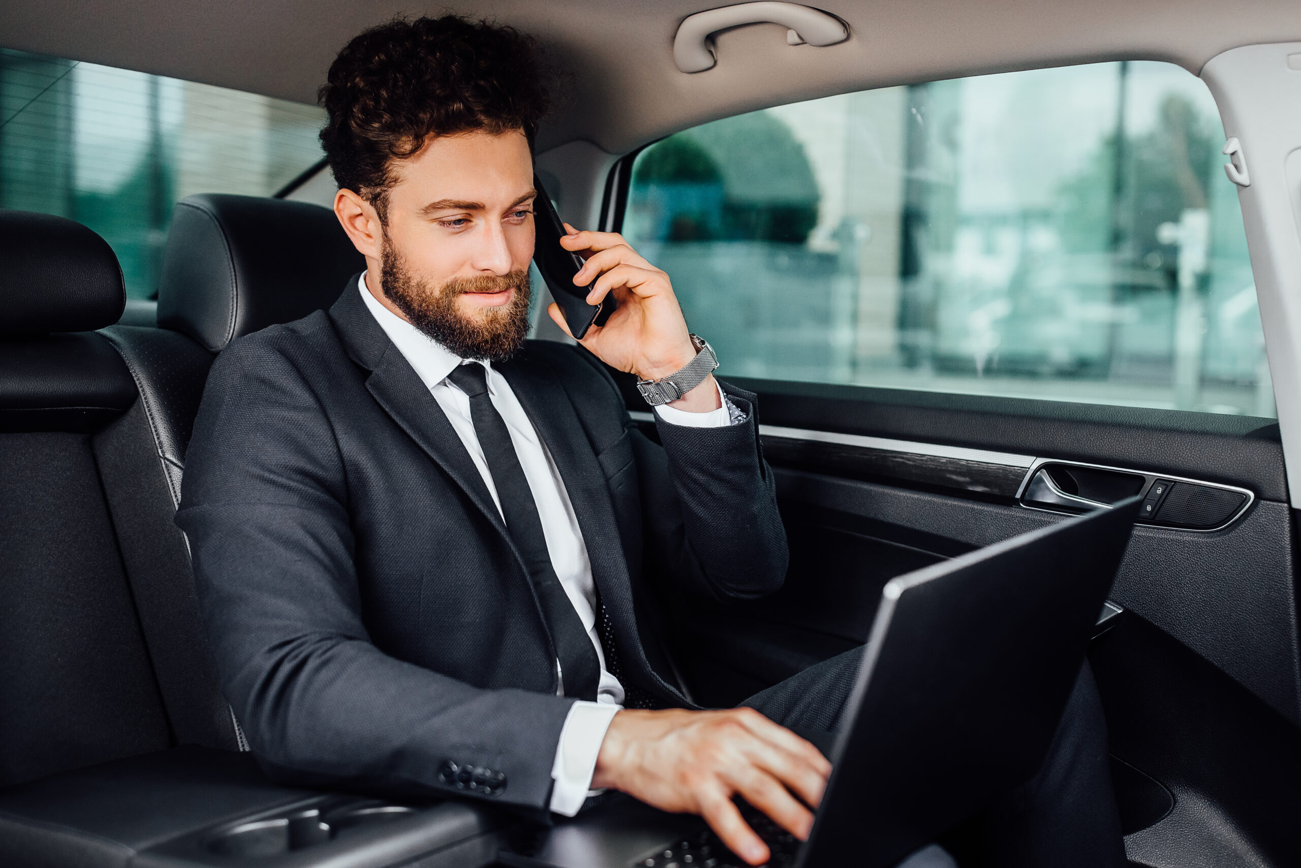 handsome bearded smiling businessman working his laptopand speaking mobile phone backseat car scaled